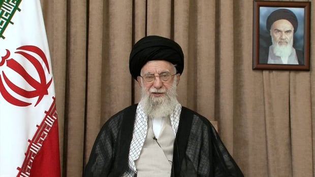 A bearded bespectacled man wearing a turban and robes is shown seated in a room.