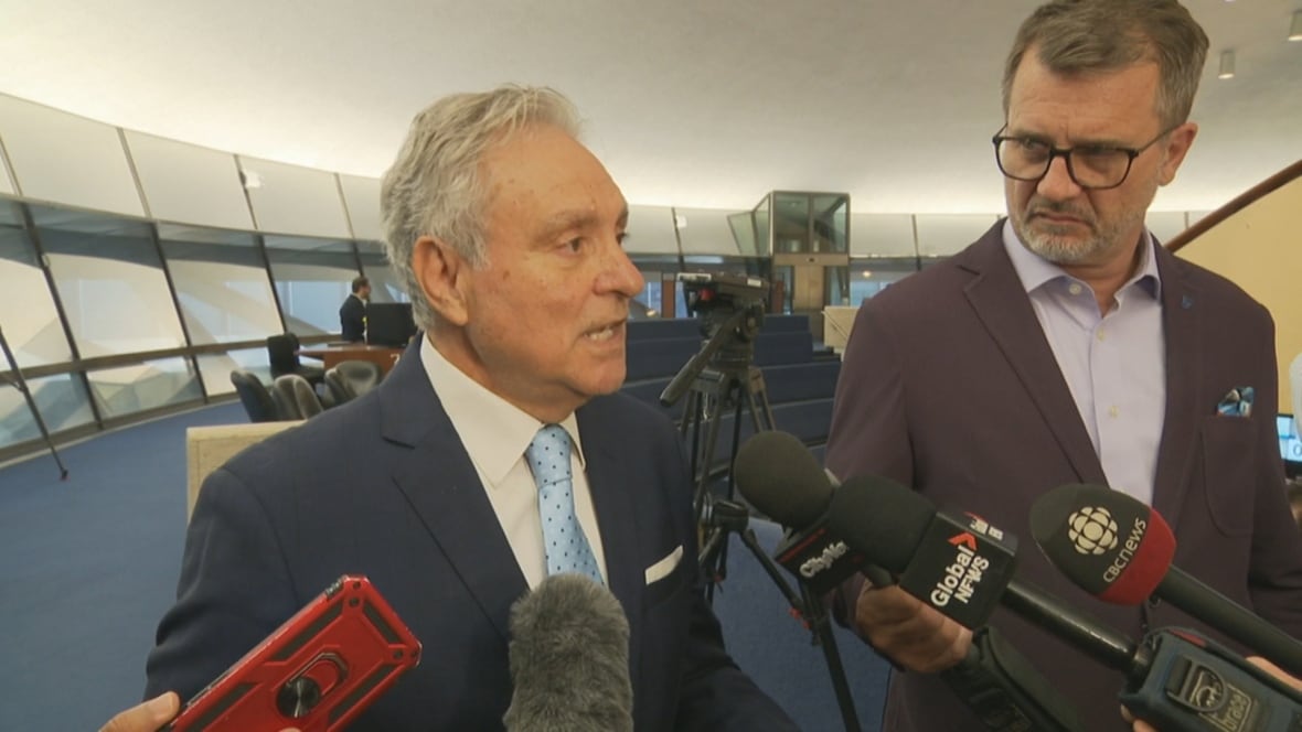 A middle-aged man with grey hair in a suit talks to reporters in Toronto City Hall