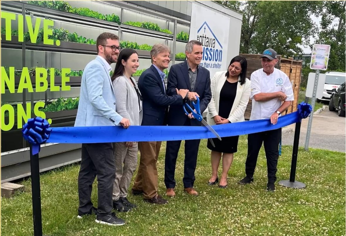 Four men and two women stand with a giant pair of scissors cutting a blue ribbon in front of a ship storage container.