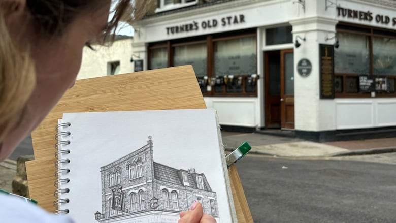 A woman with brown hair is pictured from behind sketching the exterior of the pub right in front of her.