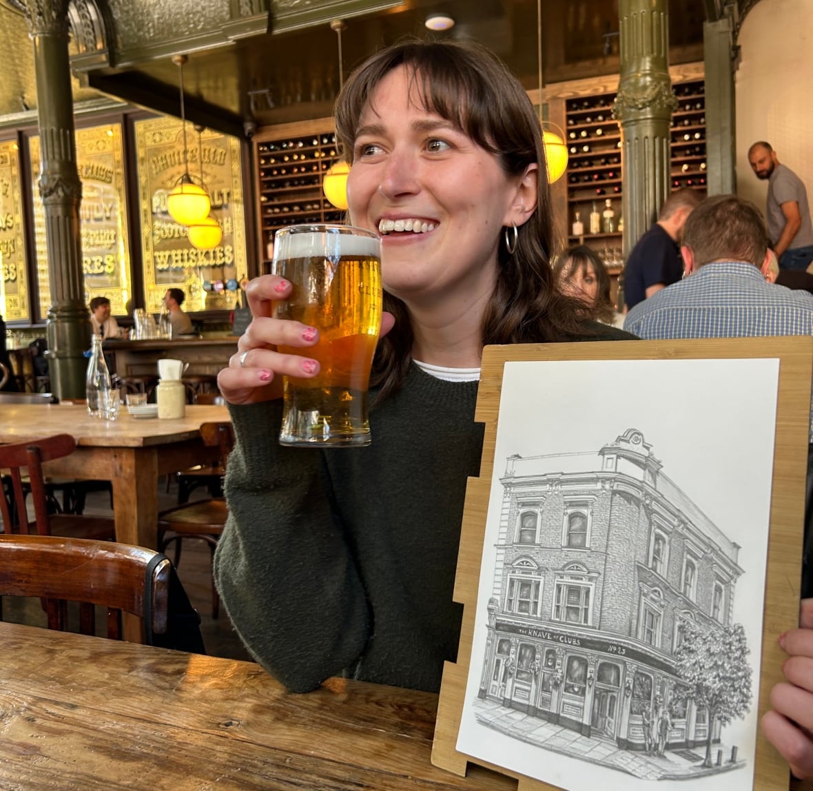 A woman smiles and sips a pint of beer in a pub while holding up a drawing of the establishment's exterior. 