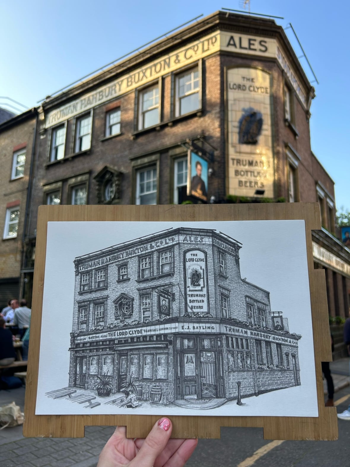 A hand holds up a pencil drawing of a corner pub in London in front of that same pub.