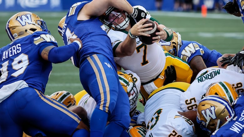 A ball carrier in white extends the football while being tackled by opponents in a pile of players at the goal line.