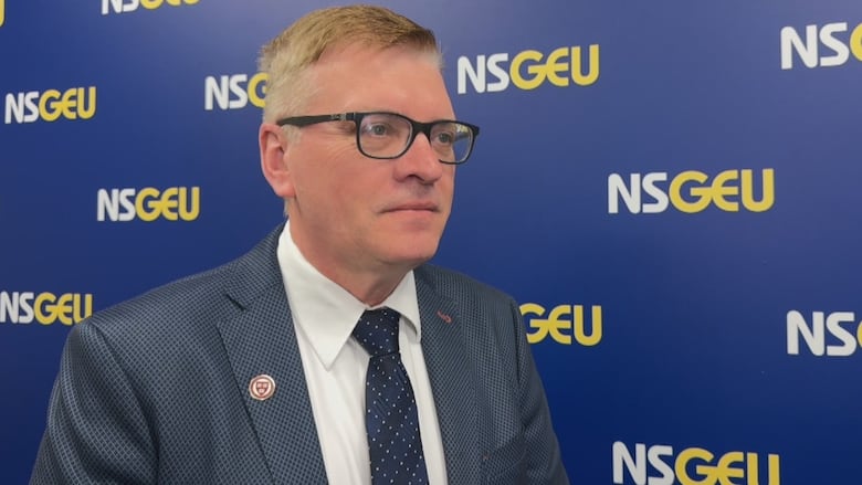 A man in a suit and tie with glasses stands in front of a wall with NSGEU written on it over and over.