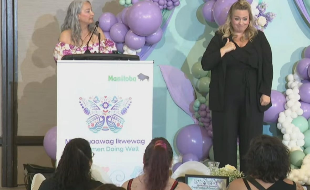 A woman speaking in front of a podium. She's looking toward another woman who is standing to her right, signing.