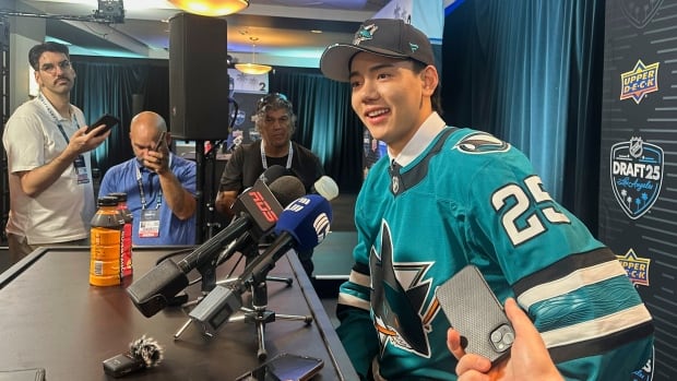 A male hockey player, wearing team jersey and hat, sits in front of an array of microphones as he answers questions from media.