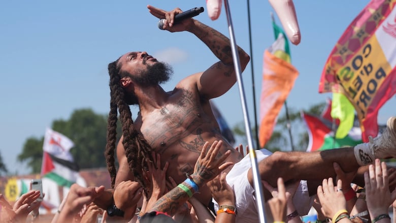A shirtless man with twisted braids, wearing white shorts, holds a microphone and looks skyward as he crowdsurfs backwards at an outdoor concert.