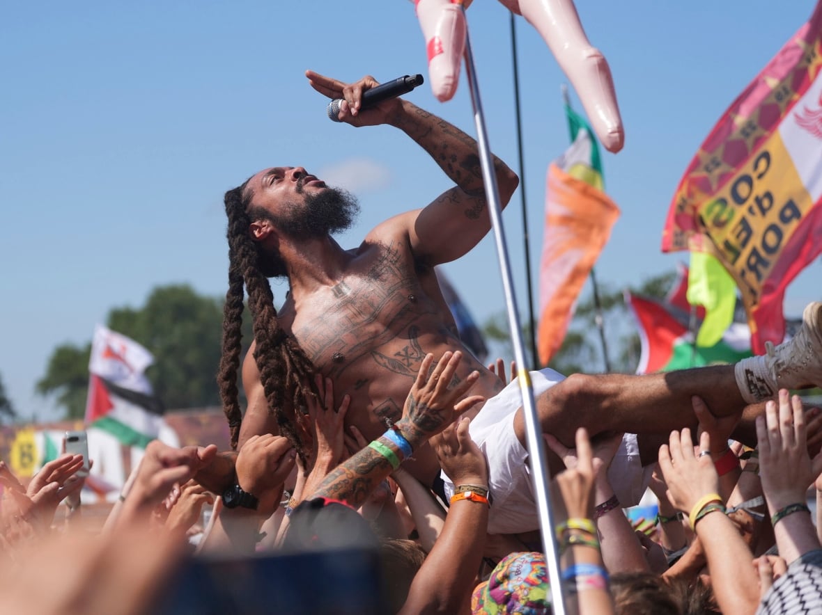 A shirtless man with twisted braids, wearing white shorts, holds a microphone and looks skyward as he crowdsurfs backwards at an outdoor concert.