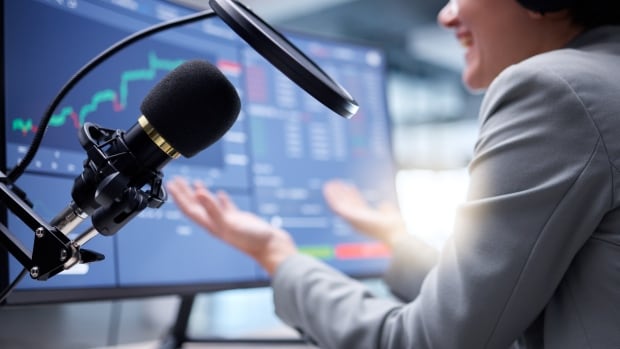 A woman is sitting in front of a computer screen with graphs on it while speaking into a podcast microphone.