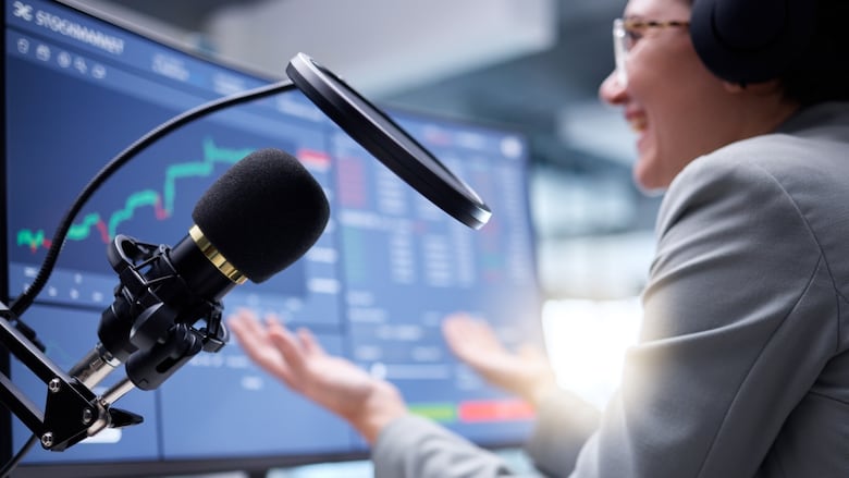 A woman is sitting in front of a computer screen with graphs on it while speaking into a podcast microphone. 
