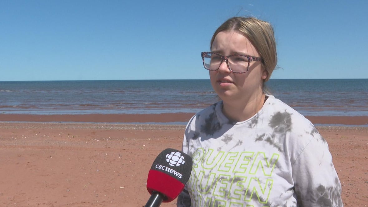 A woman in a white and gray tie dye shirt, wearing glasses talks into a CBC News microphone. She is standing on a beach and the horizon can be seen behind her.