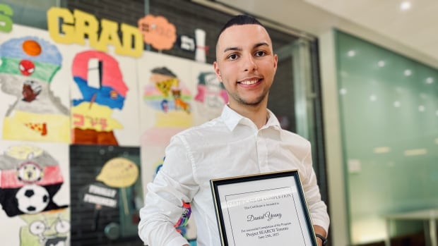 A smiling young man in a white shirt and dark pants holds a framed certificate in front of a wall decorated with brightly coloured silhouette drawings and the word GRAD.
