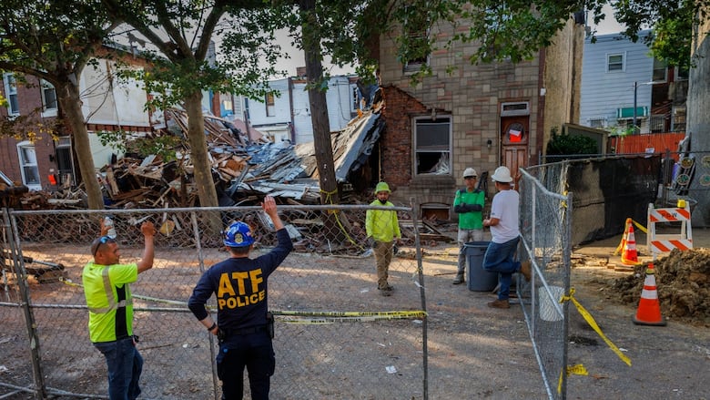 People stand around a temporary chain-link fence set up around several homes levelled or damaged by an explosion.