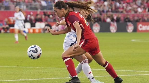 A female soccer player kicks the ball past a defender.