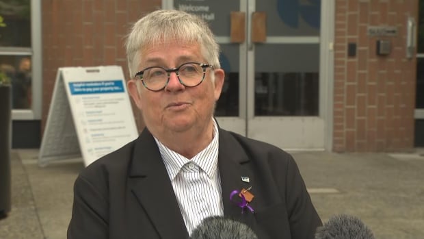 A woman wearing round glasses with short grey hair speaks into a microphone.