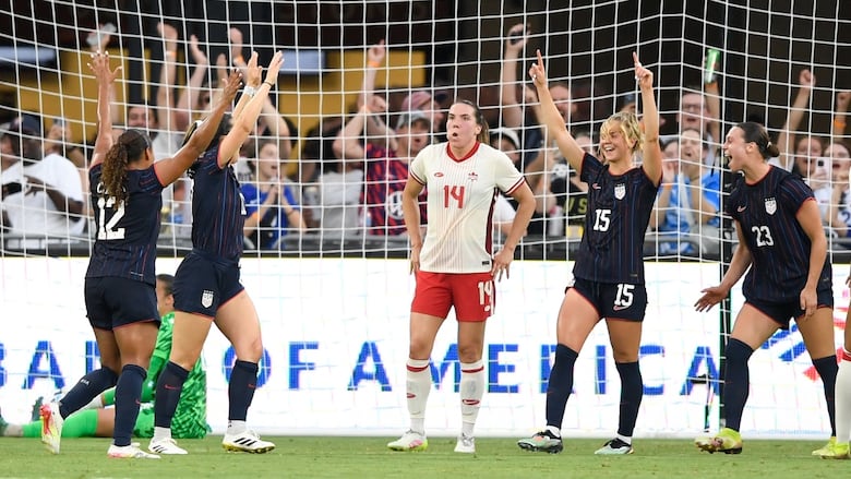 Four women's soccer players wearing dark blue kits raise thier arms in celebration in front of a soccer goalmouth, while a woman in a white kit stands in dejection in the centre.