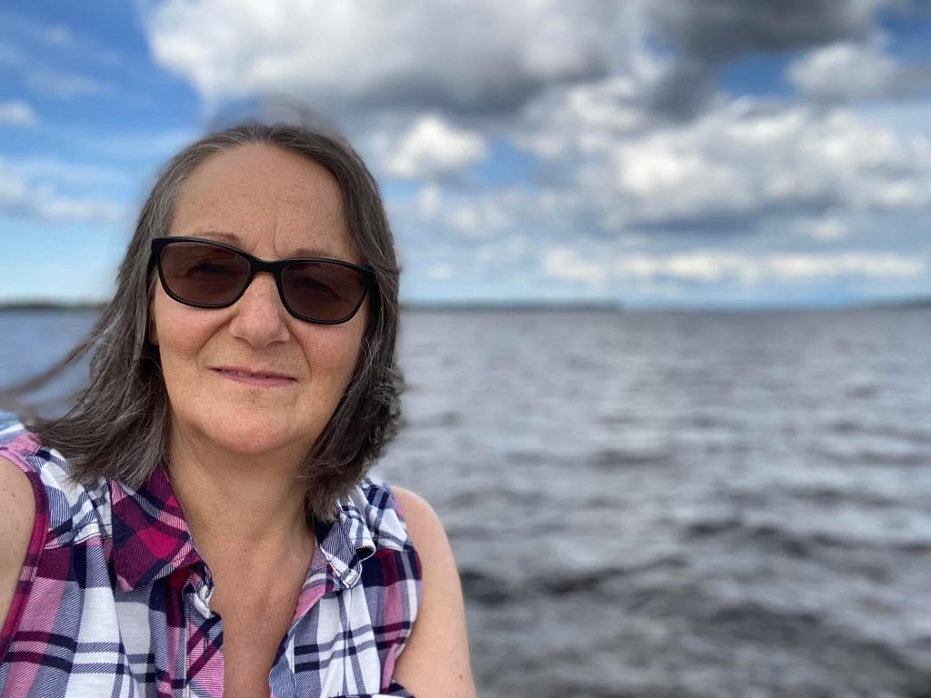 Portrait of a smiling Inuk woman wearing a plaid outfit with the ocean as a background.