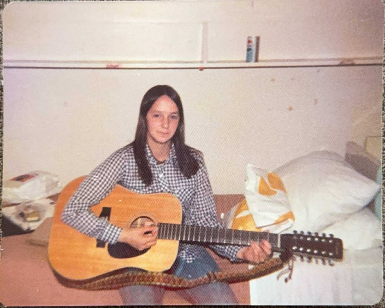 Young woman sits on a bed in a dorm room holding a guitar.