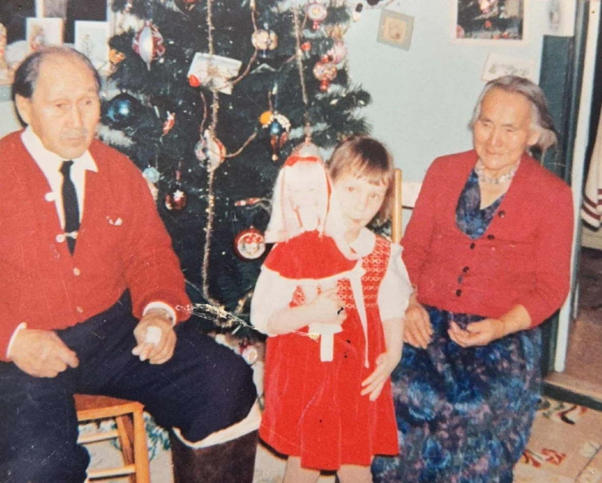 A young girl holds a doll in front of a Christmas tree with her two grandparents on each side of her.