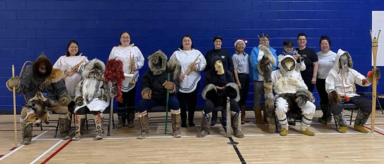 A group photo of people standing with brass instruments and people wearing furs and skins sitting on chair in a gymnasium.