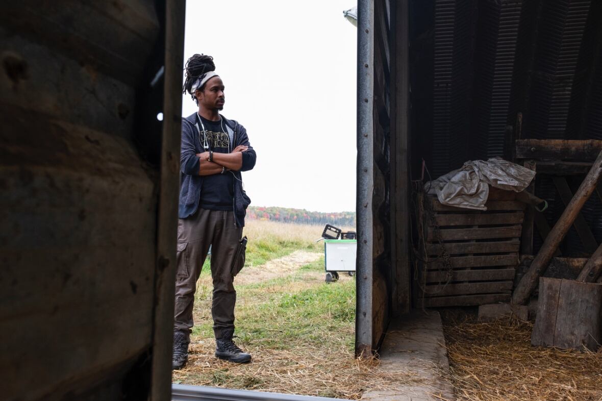A man with long dark hair tied up on his head, stands outside a barn door with his arms crossed.
