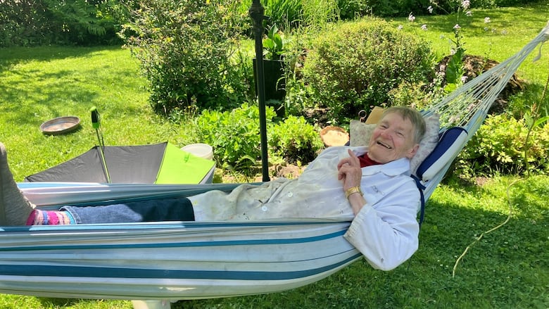 An older woman lying in a hammock in a painter's smock smiling. 
