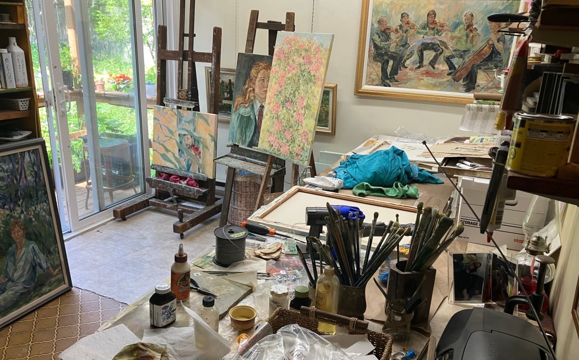Painting supplies on a desk and many easels/paintings in the background.