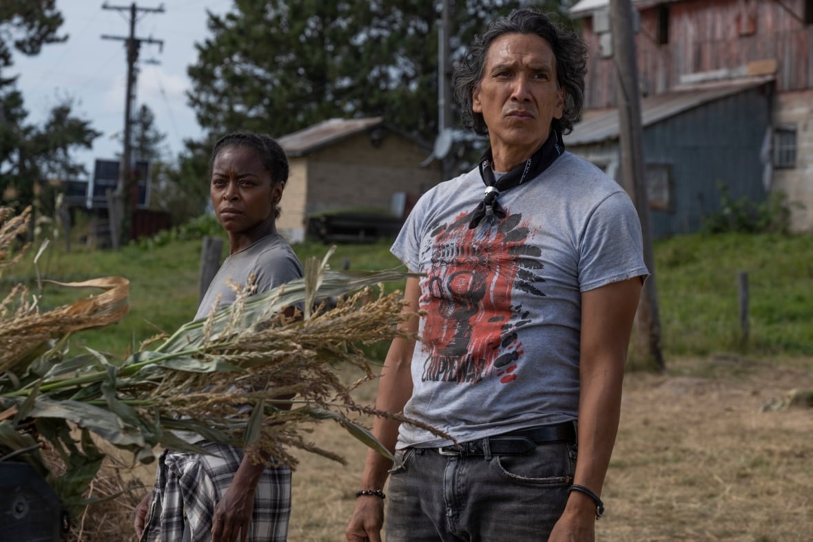 Standing on a farm property, a man with medium-length hair and handkerchief tied around his neck and a woman with dark braided hair stare into the distance.