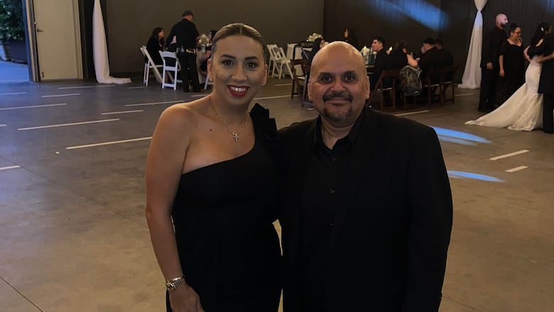 A man and a woman in black formal wear pose for a photo at a wedding.