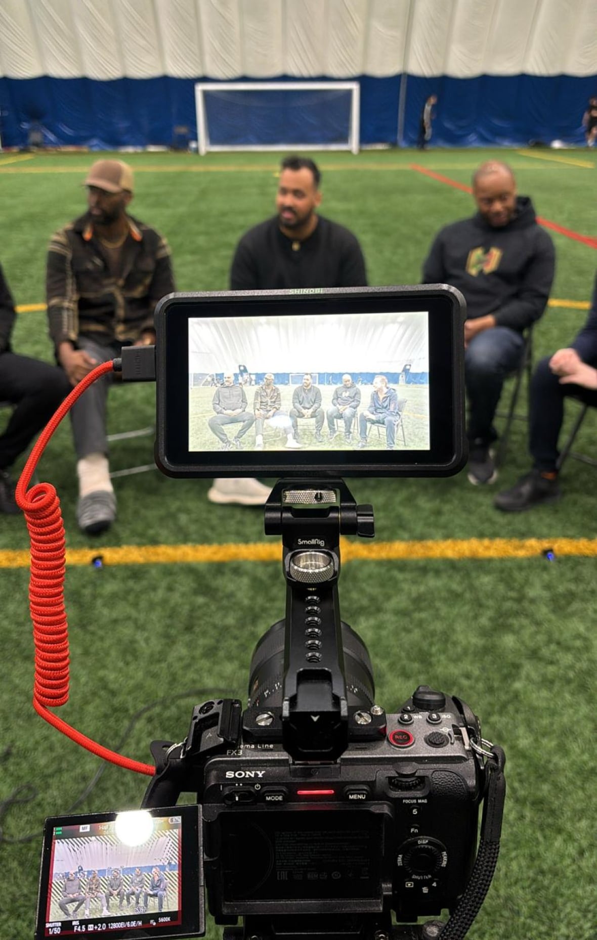 A photo of three men on a soccer field with a video camera in the foreground.
