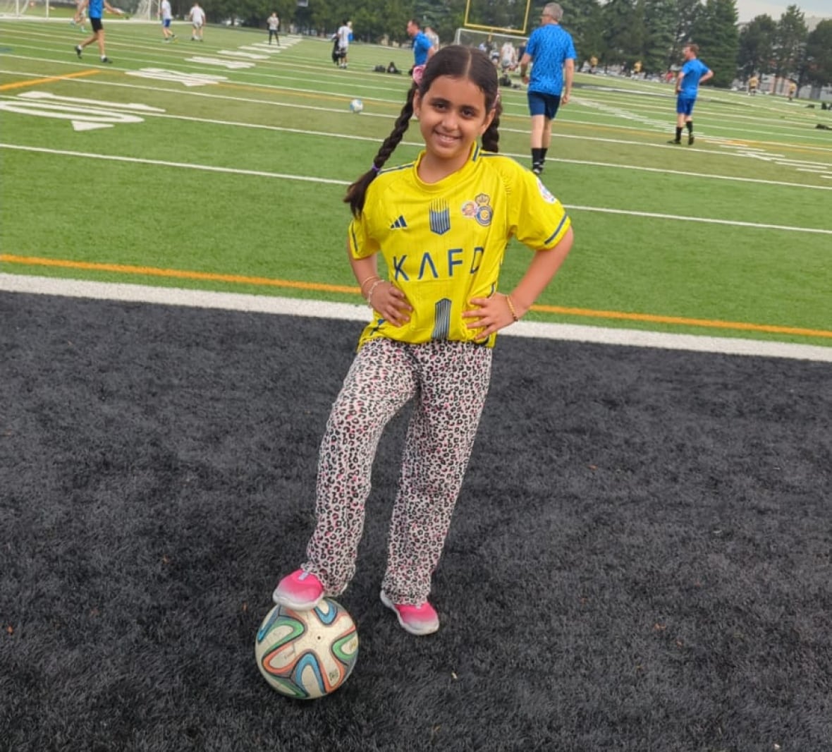 A young girl in braids stands with a soccer ball on a field.