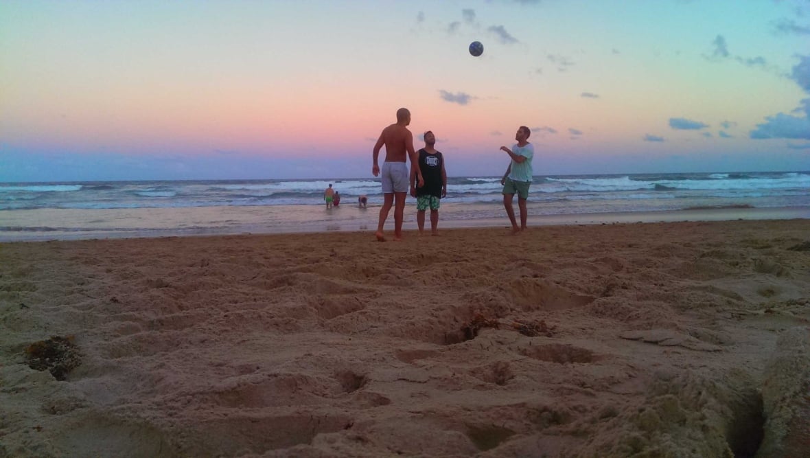 A picture of three men playing soccer at sunset on a beach.