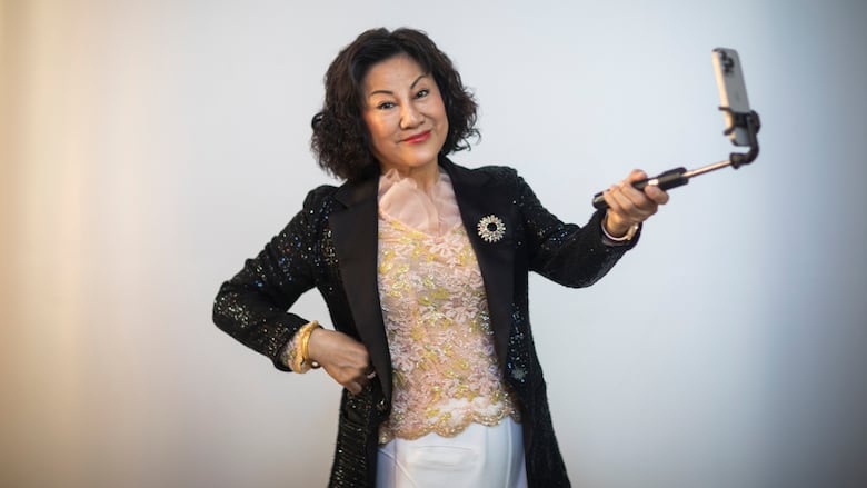 An East Asian woman with a dark bob haircut smiles while holding a selfie stick. She wears a sparkly pink and green top with a sparkly black blazer over top.