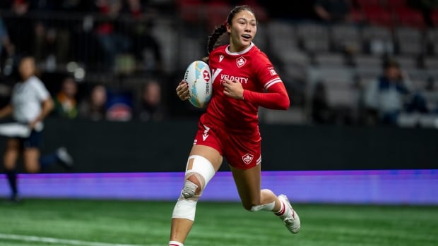 A female rugby player runs with the ball tucked by her side.