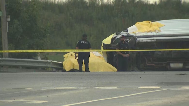 2 police officers stand by a flipped white van on a highway on a grey day. The scene is taped off