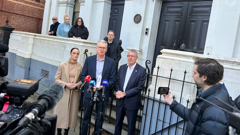 A group of people stand outside a building with microphones pointed at them.