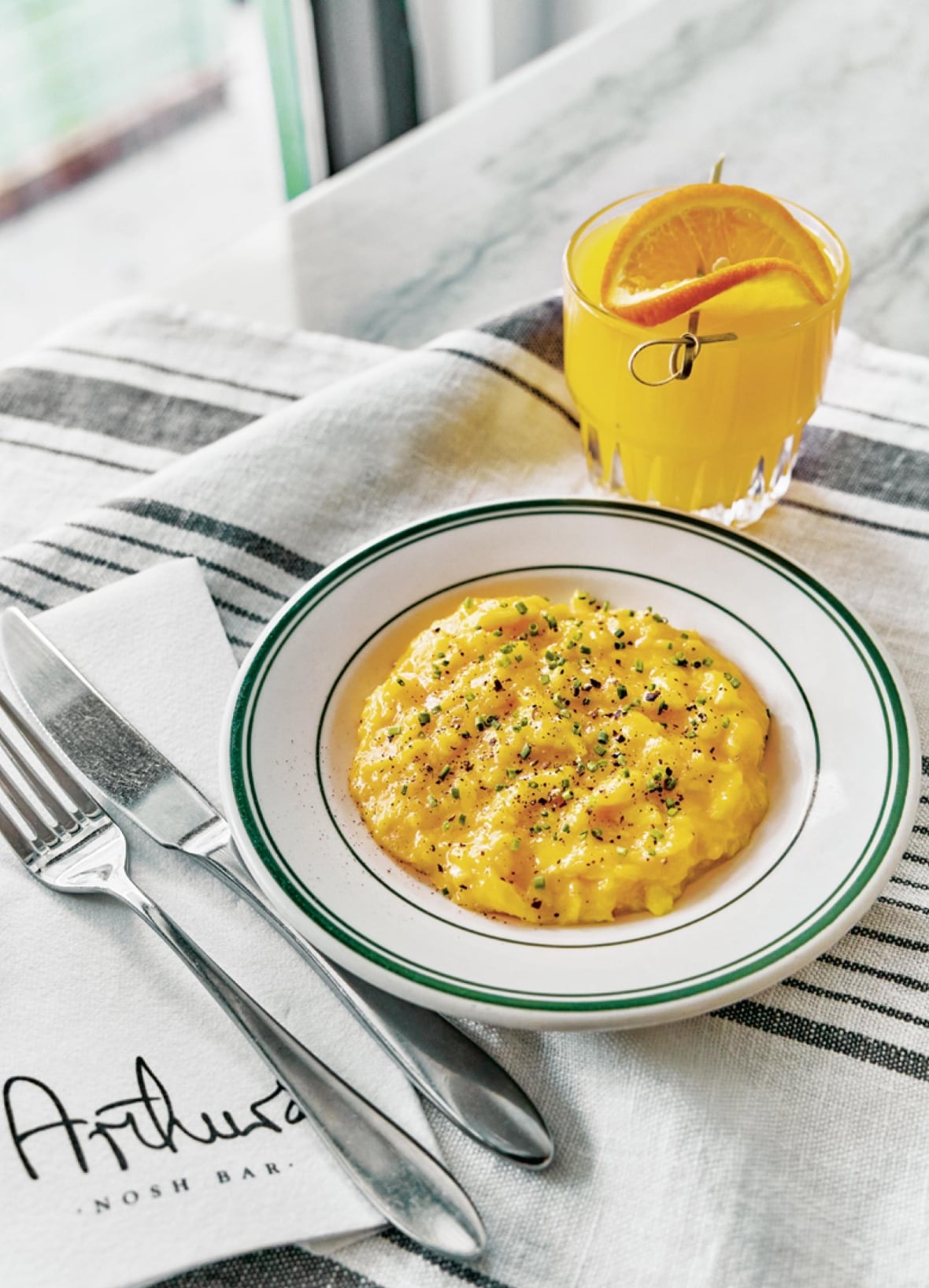A plate of scrambled eggs topped with black pepper and chives, with a glass of orange juice and fork and knife next to the plate.