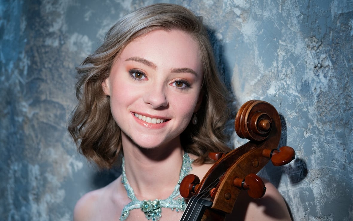 Luka Coetzee poses with her cello before a blue background.