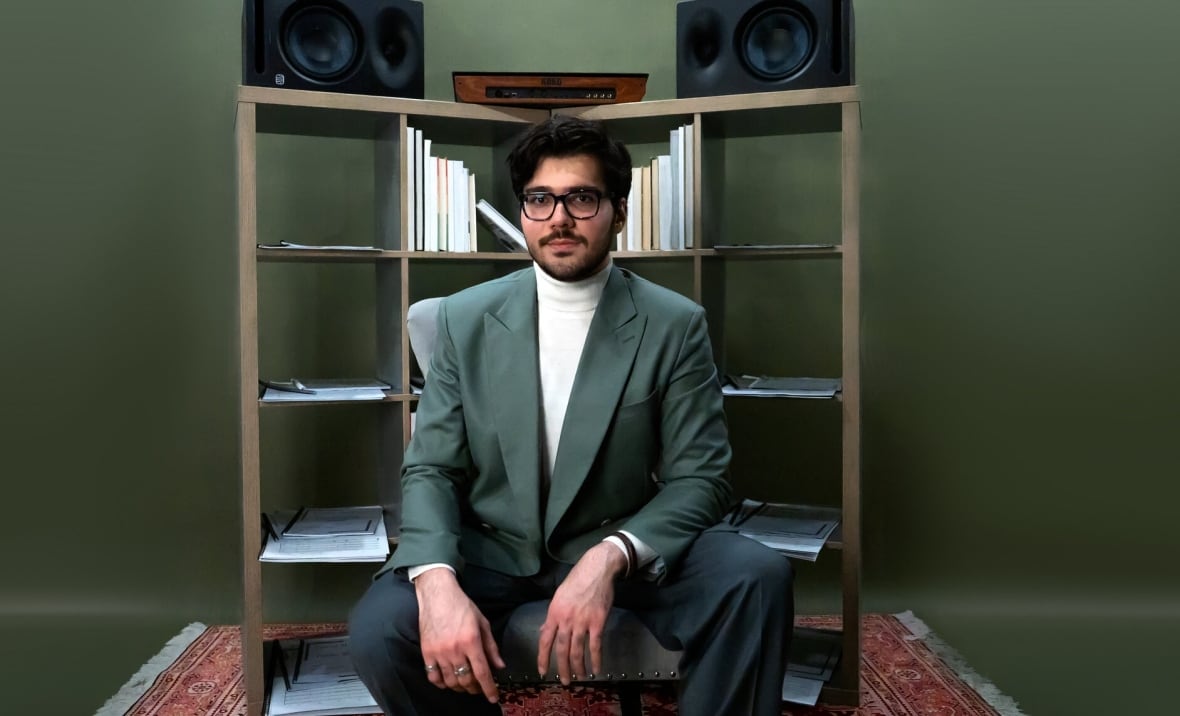 Composer Danial Sheibani is seated in his studio with a bookcase and stereo speakers in the background.