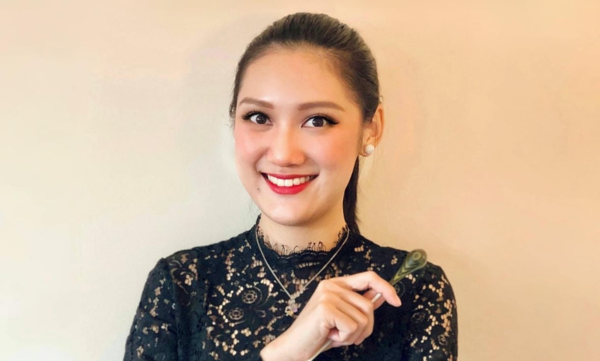 Conductor Meijun Chen, wearing a black lace dress, poses against a peach-coloured backdrop, holding a baton.