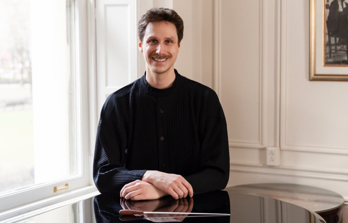 Wearing a black shirt, conductor Benoit Gauthier sits behind the surface of a shiny grand piano with his baton placed next to his hands.
