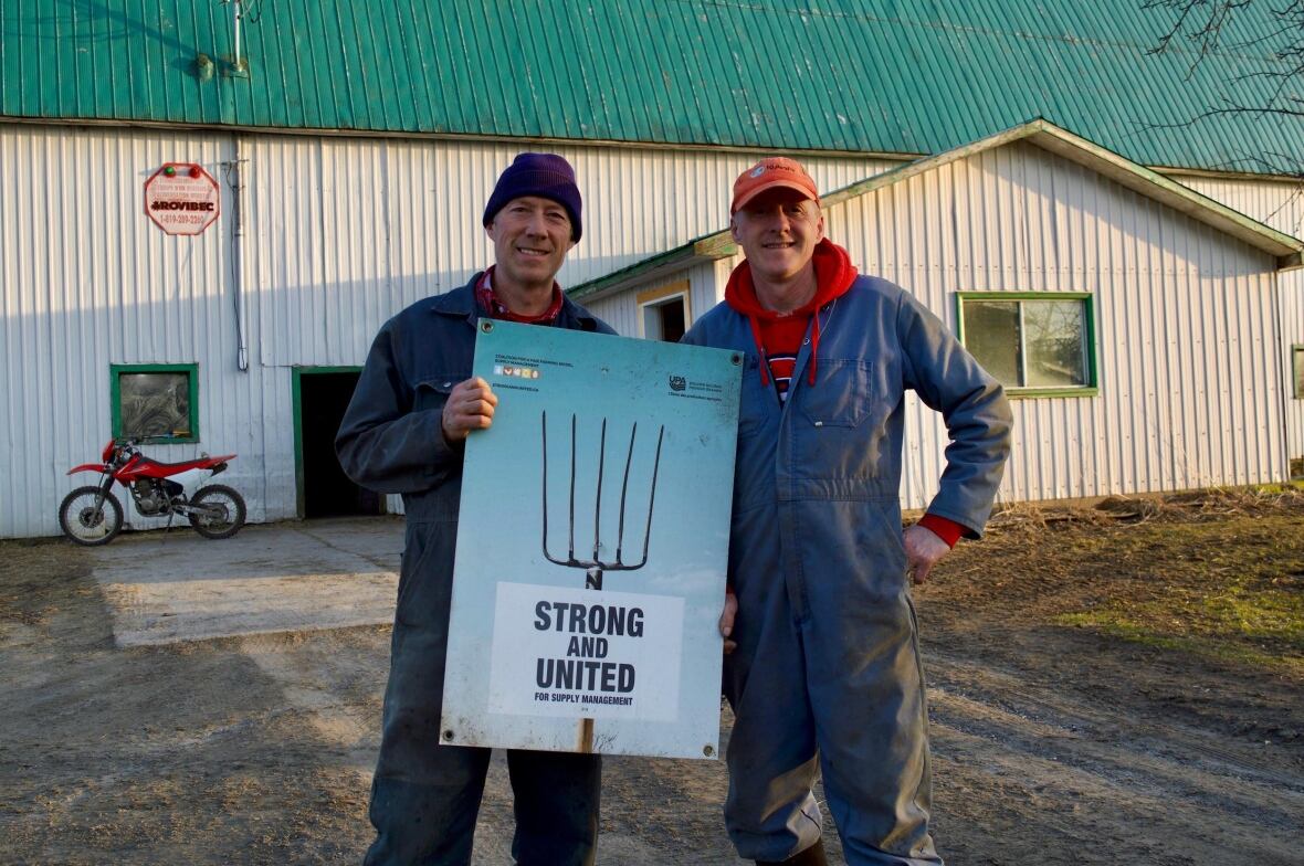Two men holding a sign while standing by a barn. 