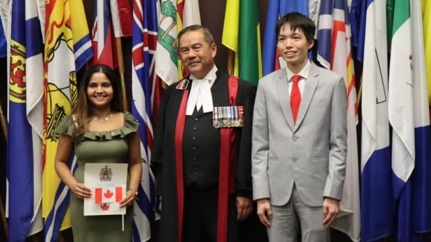 A man wearing formal robes standing beside a smiling woman holding an official document and a man dressed in a suit, with flags in the background.