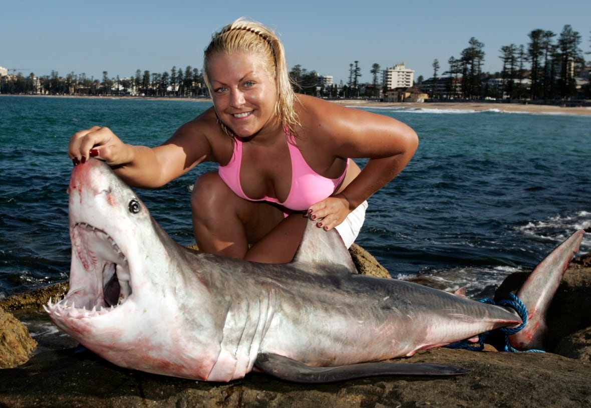 A woman in a bright pink bikini top holds open the jaw of a shark near a beach.