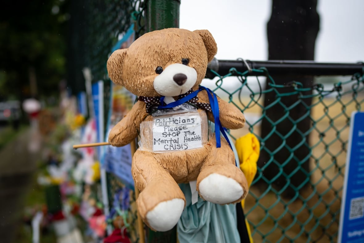 A teddy bear is hung on a chain-link fence to memorialize the lives lost at a Lapu Lapu block party on April 26, 2025. It has a sign reading 'Politicans please stop the mental health crisis'.