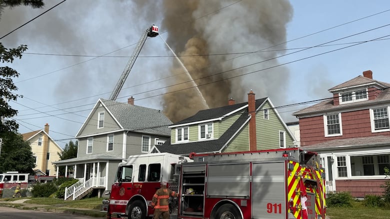 Water from an aerial ladder is sprayed on smoke coming from behind two residential homes. On the street, a firefighter stands by a parked firetruck.