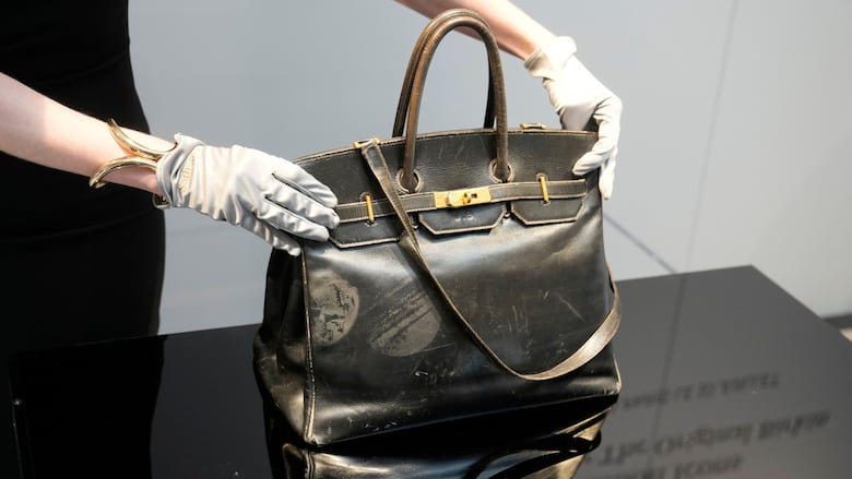 A woman's white-gloved hands clasp  a achromatic  leather handbag upright.