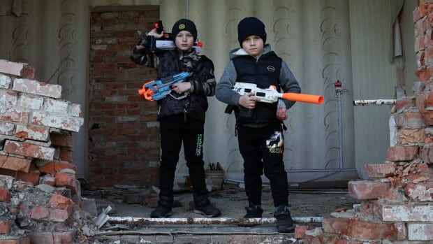 Two boys stand in a building near a brick wall that was been destroyed holding toy guns.