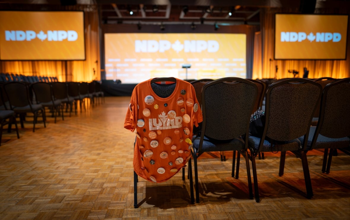 An orange shirt covered in buttons lays drapped over a chair in a convention hall. Big orange signs in the background read "NDP."