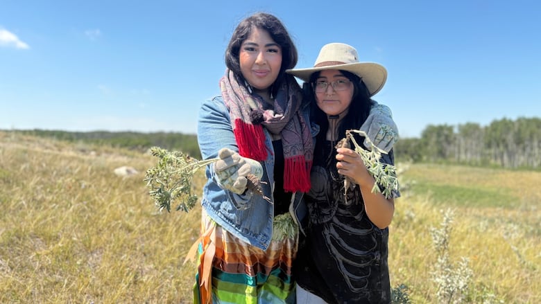 A woman in a jean jacket and ribbon skirt poses with her teenaged sibling, who is wearing a dark t-shirt and light sweatpants with a cowboy hat, while holding each a wild turnip plant, which is a flower that has a big bulb at the bottom.
